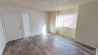 This single-floor space offers a clean, functional layout with a bright and open feel. The main living area is finished with durable wood-look laminate flooring in a warm gray-brown tone, providing a modern yet understated aesthetic that pairs well with the neutral, off-white walls throughout. Natural light enters generously through multiple windows fitted with horizontal blinds, allowing for adjustable brightness and privacy. A glazed side panel beside the entry door further enhances the flow of natural light into the space. The textured popcorn ceiling adds a classic touch overhead. The room's open layout and light color palette create an airy atmosphere, making the space feel welcoming and versatile. Electrical outlets are conveniently positioned along the walls, and the clean baseboard trim provides a neat, finished look throughout. The overall design is straightforward and practical, offering a neutral canvas well-suited for a variety of furnishing styles and personal tastes.