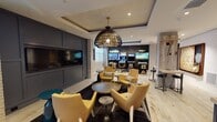 This thoughtfully designed single-floor amenity space in Woodbridge, VA, blends contemporary style with multi-functional entertainment areas, creating a dynamic environment suited for socializing and recreation. The open layout connects several distinct zones, each with its own character while maintaining a cohesive design language throughout.

The main lounge area centers around a striking woven pendant light suspended from a coffered ceiling, illuminating a seating arrangement of mustard-yellow accent chairs grouped around a dark round coffee table. A large-format TV is recessed into a slate-gray paneled feature wall, flanked by whitewashed brick columns that add textural contrast. A dartboard and high windows with frosted glass panels allow diffused natural light to fill the space, enhancing its open feel.

Adjacent to the lounge, a dedicated game room houses classic arcade machines and pinball tables alongside wall-mounted screens, offering a lively entertainment corner. A bar-height counter with black stools provides casual seating nearby.

The dining area features natural wood communal tables with bench seating beneath a cluster of mixed pendant lights, accompanied by a kitchenette with a sink and under-counter refrigerator. Light wood-look flooring and white tile run throughout, tying the entire space together with a clean, modern aesthetic.