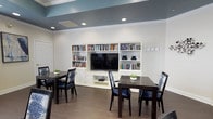 Located at 8101 Fonthill Court in Langhorne, Pennsylvania, this single-floor community space is thoughtfully designed to offer both comfort and functionality. The room's most prominent feature is a large, built-in white entertainment and bookcase wall unit, which houses a wide-screen television at its center, flanked by generously stocked bookshelves filled with a diverse collection of reading material. Cabinet storage at the base of the unit adds to its practicality. The ceiling draws the eye upward with a striking two-tone design — crisp white crown molding frames a bold teal-blue recessed section, giving the room a polished, contemporary character. Recessed lighting is evenly distributed throughout, ensuring the space is well-lit and inviting. Dark hardwood-style flooring grounds the room with warmth and contrast against the bright white walls. Multiple dark wood dining tables with cushioned blue-accented chairs are arranged throughout the space, making it suitable for casual gatherings, dining, or small group activities. A row of wall-mounted hooks near the entrance adds a convenient organizational touch. Overall, the space strikes a balance between a relaxed lounge atmosphere and a functional communal gathering area, ideal for socializing and leisure.