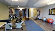 This fitness center offers a thoughtfully arranged workout space designed to accommodate a range of exercise needs. The room features a combination of hard flooring and area rugs, providing defined zones for different types of equipment. Cardio machines, including a recumbent bike and an upright exercise bike, are positioned to allow comfortable use with ample space between them. A multi-station strength training machine anchors one side of the room, offering versatile resistance training options, while a stability ball adds a functional training element nearby. Recessed lighting throughout the ceiling, complemented by pendant fixtures, ensures the space is well-lit and inviting. The walls are painted in a two-tone scheme of cool blue and warm yellow-green, giving the room a modern, energizing atmosphere. Structural columns are integrated cleanly into the layout, and two doors provide convenient access points. The overall design of the space prioritizes functionality and ease of movement, making it a practical and accessible fitness facility suitable for a variety of users and workout routines.