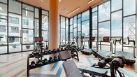 This fitness center is a thoughtfully designed amenity space that combines functionality with striking visual appeal. The layout is defined by expansive floor-to-ceiling windows framed in dark steel, flooding the interior with abundant natural light and offering impressive views of the surrounding city skyline and an adjacent outdoor terrace with seating areas. The towering glass walls create an open, airy atmosphere that makes the space feel connected to its urban surroundings.

The flooring throughout is a standout design element — a bold chevron-patterned hardwood in a mix of natural and blue-toned planks that adds a contemporary, energetic character to the space. Warm terracotta and blush-toned walls complement the natural wood tones, while recessed lighting and linear ceiling fixtures ensure well-lit workouts at any time of day.

The fitness area is comprehensively equipped with cardio machines including treadmills and ellipticals positioned along the glass walls to maximize the views, as well as a full dumbbell rack, adjustable benches, functional training equipment, kettlebells, medicine balls, and resistance tools. A dedicated stretching and open training area provides additional versatility. Direct access to the outdoor terrace further extends the usable fitness space and enhances the overall experience.