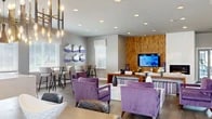 Located at 25 Pittston Cir in Owings Mills, Maryland, this single-floor community amenity space offers a well-appointed and thoughtfully designed environment spanning a generous footprint. The space is divided into distinct functional areas, each reflecting a cohesive modern aesthetic anchored by a bold purple and neutral gray color scheme throughout.

The main lounge area is an inviting, open-concept gathering space with soaring vaulted ceilings, recessed lighting, and wood-look tile flooring laid in a herringbone pattern. A striking natural wood accent wall frames a wall-mounted flat-screen TV, while a sleek built-in linear fireplace adds warmth and ambiance. Plush purple velvet seating arrangements, vibrant abstract artwork, and a stylish pendant chandelier contribute to an upscale, contemporary feel. A separate dining nook with bar-height seating and large windows allows for ample natural light.

The kitchenette features warm wood cabinetry, a bold purple subway tile backsplash, quartz countertops, and a window overlooking the outdoors — ideal for entertaining or casual use.

The fitness center is well-lit with large windows offering outdoor views, vaulted ceilings, and durable rubberized flooring. It is equipped with a variety of cardio and strength training machines. Two accessible restrooms with grab bars, lavender walls, and tile flooring round out this amenity-rich space.