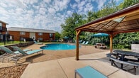 Located at 10500 Hackberry Drive in Sappington, Missouri, this outdoor amenity space centers around a generously sized, kidney-shaped swimming pool with clear blue water. The pool area is thoughtfully designed to offer a well-rounded outdoor experience, featuring a wide concrete sundeck lined with lounge chairs for relaxing under the open sky. A covered gazebo with picnic seating provides a shaded gathering spot, complemented by additional outdoor dining areas with umbrellas for sun protection. The pool is enclosed by a sturdy black iron fence, providing both safety and a defined boundary for the space. Steps lead up to a well-manicured grassy area, connecting the pool zone to the surrounding brick residential buildings, which feature private balconies overlooking the grounds. Mature trees border the perimeter, offering natural shade and greenery that enhance the overall ambiance. Pool rules signage and safety equipment are clearly posted near the entry points. The combination of the lush landscaping, open sky, and classic brick architecture creates a welcoming and functional outdoor retreat suited for warm-weather relaxation and community gatherings.