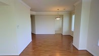 This single-floor space presents a clean, open layout with a comfortable sense of scale. The main living area is finished with warm hardwood flooring that runs consistently throughout, contributing to a cohesive and inviting atmosphere. Crown molding traces the perimeter of the ceiling, adding a refined architectural detail that elevates the overall aesthetic.

A pendant light fixture with a classic cone-shaped shade hangs centrally, serving as a focal point and providing ambient illumination to the space. Natural light enters through a window on the side wall, brightening the interior and complementing the neutral, light-toned walls.

The open floor plan allows for a natural flow between living areas, with a visible transition into an adjoining space toward the rear. Tiled flooring in the background area suggests a functional transition zone, possibly leading to a kitchen or entryway. The layout offers flexibility for various furniture arrangements.

Overall, the space conveys a well-maintained, move-in ready character, with traditional detailing such as the crown molding balanced by straightforward, practical design choices suited to everyday living.