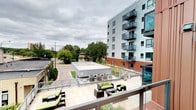 This single-level outdoor terrace is part of a modern residential development characterized by a striking contemporary architectural design. The building exterior features a combination of light grey paneling and warm wood-toned cladding, creating a visually dynamic facade. Individual balconies with glass railings extend from the upper floors, reinforcing the building's modern aesthetic.

The terrace itself is finished with large-format light-toned pavers, providing a clean, polished surface throughout the space. The outdoor area is furnished with multiple seating arrangements, including dark wicker lounge sets with green cushions, creating comfortable gathering spaces suited for relaxation or socializing. Additional seating is visible along the perimeter, maximizing use of the terrace footprint.

HVAC equipment is neatly positioned within a designated section of the terrace, maintaining functionality while preserving the overall aesthetic. The terrace overlooks a street-level courtyard and surrounding tree canopy, offering pleasant greenery views. The elevated vantage point provides sightlines across neighboring rooftops and into the broader urban streetscape, contributing to an open, airy ambiance throughout the outdoor space.