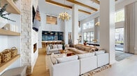Located at 2001 E Fourth St in Fort Worth, TX, this single-floor clubhouse delivers an impressive blend of modern design and warm, inviting character across a generously sized open-plan layout. The living area immediately commands attention with its dramatic double-height ceilings adorned with exposed wood beams and a statement multi-globe brass chandelier. Wide-plank hardwood flooring flows throughout, grounding the bright, airy interior. A floor-to-ceiling stacked stone fireplace anchors one wall of the living area, which is furnished with oversized cream sectional sofas centered around a solid wood coffee table, creating an ideal social gathering space. Large windows on multiple walls allow abundant natural light and offer views of the surrounding Fort Worth streetscape. The kitchen features slate-blue cabinetry, a spacious white quartz island with gold bar stools, herringbone tile flooring, and bold cone-shaped brass pendant lights. A cozy dining nook is tucked nearby, finished in rich slate tones with built-in shelving, a leather banquette, and a round walnut dining table. Steel-framed glass partitions provide subtle separation between zones while preserving the open, connected feel of the entire clubhouse.