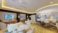 This elegantly designed single-floor amenity lounge offers a refined and welcoming environment characterized by a cohesive contemporary aesthetic. The space is anchored by a coffered ceiling with warm amber cove lighting that runs along the perimeter, complemented by recessed downlights that illuminate the room evenly throughout.

The main lounge area features multiple comfortable seating arrangements with crisp white upholstered sofas, accent chairs, and glass coffee tables arranged atop layered rugs, creating intimate gathering zones. Twin flat-screen televisions are mounted within dark wood-paneled wall niches accented with gold trim detailing and sleek wall sconces, offering both visual focal points and functional media access. A built-in workstation niche provides a printer and dedicated workspace.

A curved bar counter with white high-gloss bar stools sits adjacent to a refreshment station, which includes a wall-mounted television, light wood cabinetry, quartz countertops, a built-in appliance, and snack and beverage service. Marble-topped dining tables with modern chairs offer additional seating throughout.

Large floor-to-ceiling glass doors along one wall flood the interior with natural light and provide direct views and access to an outdoor terrace. The light wood-toned flooring, neutral color palette, and carefully curated greenery and artwork contribute to the space's polished, hospitality-inspired ambiance.