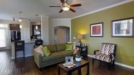 This single-floor residence showcases a thoughtfully designed open-concept layout that blends comfort and style. The living area is a standout feature, highlighted by rich dark hardwood flooring, elegant crown molding, and a ceiling fan that adds both function and character. An accent wall painted in a warm olive green creates a striking focal point, complementing the earthy tones found throughout the space. An arched passageway adds an architectural touch, lending a sense of depth and elegance to the layout.

The living room is spacious and well-lit by ambient lighting, centered around a plush olive-green sofa adorned with decorative pillows. A dark wood coffee table and coordinating side table anchor the seating area, while framed wall art and curated décor contribute to the polished aesthetic.

The space flows seamlessly into the kitchen, which is visible from the living area and features dark cabinetry, stainless steel appliances, and a breakfast bar with pendant lighting overhead. The open sightlines between rooms enhance the sense of space and create an inviting, connected atmosphere ideal for both everyday living and entertaining.