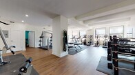 This comprehensive fitness center is thoughtfully designed across a single floor, offering a wide range of exercise zones to accommodate diverse workout needs. The main workout area features warm hardwood flooring, white walls, and a coffered ceiling with structural beams that lend an architectural character to the space. Large windows line the cardio zone, flooding the area with natural light and offering views of the surrounding cityscape, creating an energizing environment for exercise. This section houses multiple treadmills, elliptical machines, and stationary bikes arranged in an orderly row. A dedicated strength training area features a multi-station cable machine, adjustable benches, and wall-mounted mirrors that visually expand the space. Resistance bands and accessories are neatly organized on wall-mounted racks. A vibrant teal-painted spin room provides an intimate, high-energy setting with spin bikes and a full-length mirror. Additional teal-accented rooms offer space for free weights, kettlebells, stability balls, and stretching, each equipped with mirrors and recessed lighting. Two full bathrooms, finished with white subway tile, marble-look flooring, glass-enclosed soaking tubs, and chrome fixtures, provide polished post-workout facilities. Overall, the facility balances functionality with a clean, modern aesthetic.
