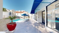Located at 333 Andover Dr in Burbank, California, this outdoor amenity space is designed to maximize the region's abundant sunshine and mild climate. The centerpiece is a large, crystal-clear swimming pool surrounded by a generously sized concrete sundeck furnished with multiple blue lounge chairs, perfect for relaxing under the Southern California sun. A separate spa with tiled surround provides an additional area for leisure, positioned alongside shaded outdoor dining with a blue umbrella-covered table and chairs. Vibrant red ceramic planters and tropical greenery add pops of color and a resort-like aesthetic to the space. A striking blue canopy awning marks the entrance to an adjoining room that houses fitness and recreation equipment, visible through large glass doors and windows. The buildings surrounding the pool area feature a white stucco exterior with wrought iron balconies, terracotta rooflines, and architectural detailing that lends a Mediterranean-inspired character to the complex. Mature trees frame portions of the property, offering natural shade and a sense of privacy. The overall ambiance is bright, clean, and welcoming, offering a well-rounded outdoor retreat suited for relaxation and recreation year-round.