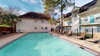This amenity space offers residents two distinct areas designed for recreation and fitness. The outdoor pool area features a generously sized, clear-water swimming pool enclosed by a black iron fence, surrounded by a brick-trimmed pool deck lined with lounge chairs. Mature pine and palm trees provide natural shade, creating a relaxed, resort-like atmosphere set against the backdrop of the surrounding residential buildings.

Adjacent to the pool, the fitness center is a bright, well-appointed space finished with neutral gray walls, herringbone-patterned flooring, and recessed lighting complemented by a ceiling fan for air circulation. Floor-to-ceiling mirrors line one wall, visually expanding the space and enhancing natural light that streams in through large glass doors leading directly to the pool deck. The facility is comprehensively equipped with multiple treadmills, elliptical machines, stationary bikes, cable and weight machines, a full dumbbell rack, medicine balls on a dedicated stand, and a free-weight bench. Wooden open shelving along one wall holds rolled towels, plants, and decorative accents, while a wall-mounted television adds entertainment during workouts. The direct pool-facing entry creates a seamless indoor-outdoor connection.