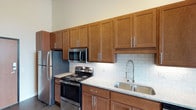This single-floor unit showcases an industrial-modern aesthetic, highlighted by polished concrete floors that run throughout the space. The kitchen is a standout feature, offering an expansive run of rich, warm-toned shaker-style cabinetry that extends to the ceiling, providing generous storage. Granite-look countertops complement the full suite of stainless steel appliances, including a refrigerator, electric range, over-the-range microwave, and dishwasher. A classic white subway tile backsplash adds a clean, timeless detail behind the cooking area, while a spring-neck faucet serves the double-basin stainless steel sink. The bedroom area benefits from a large window that frames outdoor greenery, allowing abundant natural light to fill the room. Exposed ductwork adds an industrial character to the ceiling. The full bathroom is neatly appointed with a granite-top vanity with an undermount sink, brushed nickel fixtures, a large mirror, and a tub-shower combo with tiled surround. A closet adjacent to the bathroom also houses the water heater, keeping utilities discreetly integrated within the living space. Overall, the unit presents a clean, functional, and contemporary living environment.