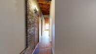 Located at 2411 E Franklin St in Richmond, VA, this 1-bedroom, 1-bathroom unit blends historic industrial character with modern conveniences. The living room immediately captures attention with its dramatic exposed brick walls, original wide-plank hardwood floors, and open wood beam ceilings fitted with visible ductwork — a hallmark of loft-style architecture. Large framed windows allow generous natural light to pour in, further enhancing the warmth of the space. A ceiling fan adds both function and style overhead.

The kitchen is efficiently designed, featuring rich cherry wood cabinetry, dark granite countertops, stainless steel appliances including a refrigerator, range, and built-in microwave, and a double stainless steel sink. A breakfast bar with countertop overhang provides casual dining space. Exposed wood beams carry through into the kitchen, maintaining the cohesive aesthetic.

The hallways are particularly striking, lined with exposed brick and illuminated by wall sconce lighting beneath the wood beam ceiling, creating an inviting passage through the unit. The bedroom continues the hardwood flooring and beam ceiling details. The bathroom offers a clean, classic finish with tile flooring, a shower/tub combo, and a warm wood vanity with ample storage.