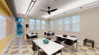 This single-story space functions as a versatile classroom or activity room, thoughtfully designed to support group learning and collaboration. The room is filled with abundant natural light streaming through multiple large windows fitted with roll-down shades, which line two walls and offer views of greenery outside. The neutral, light-toned walls enhance the sense of openness and brightness throughout the space.

Overhead, a combination of recessed linear LED lighting and a sleek black ceiling fan ensures consistent illumination and comfortable air circulation year-round. The flooring features a distinctive multi-toned geometric tile pattern in yellow, gray, and white, adding visual interest and a modern aesthetic to the room.

The furnishings consist of several rectangular white tables arranged in clusters, each accompanied by black contemporary chairs, facilitating both individual and group-oriented activities. Organizational accessories, including supply caddies on the tables, reflect the room's functional focus. A motivational poster near the entrance adds a welcoming, encouraging tone. A door on one side provides easy access, contributing to the room's practical, well-organized layout.