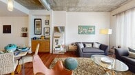 Located at 2020 Lawrence in Denver's vibrant RiNo/Curtis Park neighborhood, this single-floor residence showcases a compelling blend of industrial and contemporary design. The living area is defined by striking exposed concrete ceilings with visible ductwork, rich hardwood flooring, and large windows that frame urban city views while flooding the space with natural light. The open-concept kitchen is well-appointed with light wood cabinetry, stainless steel appliances including a refrigerator, electric range, and built-in microwave, complemented by a subway tile backsplash and a generous island with quartz countertops, a double sink, and bar seating — ideal for both cooking and casual dining. Pendant and track lighting illuminate the kitchen, adding a modern touch. The bedroom continues the industrial aesthetic with the same exposed concrete ceiling and ceiling fan, soft carpeting underfoot, and wide windows offering city views. A full bathroom features a soaking tub and shower combination with white tile surround, a vanity with wood cabinetry, and a frosted window for natural light. Throughout the residence, the interplay of raw architectural elements and warm finishes creates a distinctive, urban ambiance.