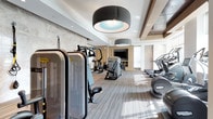 This upscale fitness center is thoughtfully designed to deliver a premium workout experience across a single, expansive floor. The space is anchored by a bright, open cardio zone lined with a row of professional treadmills and stair climbers positioned along large windows that invite abundant natural light. Decorative drum pendant lighting with coffered ceiling details above lend a refined, modern aesthetic that sets this facility apart from a standard gym.

The strength training area is equally well-equipped, featuring a multi-functional cable and power rack system, a full set of dumbbells, kettlebells in graduated weights, and medicine balls neatly organized on dedicated racks. Adjustable benches and mirrors along the wall create a functional and spacious free-weights zone. A selection of Technogym selectorized machines is strategically distributed throughout, providing options for targeted muscle training.

A flat-screen television is mounted on a stone accent wall at the far end of the space, offering entertainment during workouts. The flooring throughout consists of striped, neutral-toned carpet tiles that add warmth while providing comfort underfoot. Entry to the facility is through a glass-paneled door that connects to a bright corridor, completing the polished and professional character of this well-designed fitness space.