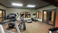 This fitness center offers a dedicated space for cardio-focused workouts, outfitted with a selection of professional-grade exercise equipment including treadmills, elliptical trainers, and a stair climber. The room is finished with durable carpeted flooring that provides comfort underfoot during exercise sessions. Large arched mirrors line the walls, visually expanding the space and allowing users to monitor their form while working out. Overhead fluorescent lighting ensures the room is well-lit throughout. The walls are painted in a neutral tone with a dark trim border detail near the ceiling, lending the space a clean and structured aesthetic. A doorway on the far side of the room provides convenient access to adjacent areas. The layout is thoughtfully arranged to allow ample circulation space between machines, ensuring a safe and comfortable workout environment. Overall, the fitness center presents a practical and functional setting suited for daily exercise routines.