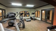 This fitness center offers a dedicated space for cardio-focused workouts, outfitted with a selection of professional-grade exercise equipment including treadmills, elliptical trainers, and a stair climber. The room is finished with durable carpeted flooring that provides comfort underfoot during exercise sessions. Large arched mirrors line the walls, visually expanding the space and allowing users to monitor their form while working out. Overhead fluorescent lighting ensures the room is well-lit throughout. The walls are painted in a neutral tone with a dark trim border detail near the ceiling, lending the space a clean and structured aesthetic. A doorway on the far side of the room provides convenient access to adjacent areas. The layout is thoughtfully arranged to allow ample circulation space between machines, ensuring a safe and comfortable workout environment. Overall, the fitness center presents a practical and functional setting suited for daily exercise routines.