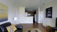 This single-floor residence offers a practical and well-appointed open-concept layout that seamlessly connects the living area to the kitchen. The living space is grounded by rich hardwood flooring that extends throughout, creating a warm and cohesive aesthetic. Crown molding adds a refined architectural detail to the space, while track lighting on the ceiling provides flexible and functional illumination.

The kitchen is positioned behind a raised half-wall peninsula, which serves both as a visual divider and a casual countertop space. Dark wood cabinetry lines the kitchen walls, offering ample storage, and is complemented by a white refrigerator and stainless appliances. A ceiling-mounted light fixture ensures the kitchen area remains bright and functional.

The living area accommodates comfortable seating, including a black leather sofa with decorative throw pillows, and is anchored by an area rug over the hardwood floor. A stylish floor lamp provides ambient lighting. Multiple doorways are visible, suggesting access to additional rooms such as a bedroom and bathroom, contributing to the overall functional flow of the space. The clean, neutral wall palette throughout keeps the interior feeling open, airy, and move-in ready.