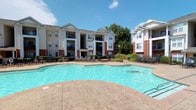 This community amenity space is situated within a well-appointed residential complex characterized by multi-story buildings featuring classic architectural details, including brick accents, neutral-toned exteriors, and private balconies. The outdoor area centers around a generously sized swimming pool with a freeform shape, surrounded by a stamped concrete deck that provides ample space for lounging. Rows of chaise lounge chairs line the poolside, offering a comfortable setting for relaxation under open skies. Mature trees and ornamental landscaping frame the perimeter, creating a pleasant natural backdrop.

The interior portion of this space functions as a business center, designed with a clean, professional aesthetic. Walls feature a two-tone color scheme — warm taupe on the upper half and deep navy blue on the lower half — separated by crisp white chair rail molding and complemented by crown molding at the ceiling. Hardwood-style flooring runs throughout, adding warmth to the space. Large windows fitted with horizontal blinds allow natural light to fill the rooms. The business center includes dedicated workstations equipped with desktop computers and ergonomic seating, as well as a utility area housing a multifunction printer, making it a fully functional workspace for residents.