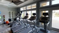 This fitness center offers a sleek, modern environment designed for effective and enjoyable workouts. The space is anchored by a row of commercial-grade cardio machines, including treadmills and ellipticals, each equipped with individual digital display screens for entertainment and performance tracking. Large windows line the walls, flooding the room with abundant natural light while offering outdoor views that create an open, energizing atmosphere. The interior is finished in a sophisticated color palette of deep gray-green tones, complemented by tray ceiling details and a ceiling fan that enhances air circulation throughout the space. Built-in shelving units along one wall provide convenient storage, adding both function and structure to the layout. The flooring is tastefully tiled, contributing to the clean, polished aesthetic of the room. The overall ambiance strikes a balance between modern design and practical utility, making it a welcoming destination for daily fitness routines. Whether used for a brisk morning run or a focused cardio session, this fitness center is thoughtfully appointed to meet the needs of its users in a comfortable and visually appealing setting.