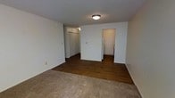 This single-floor space offers a practical and straightforward layout designed for comfortable everyday living. The main living area features a combination of plush carpet and dark hardwood-style flooring, creating a clear visual distinction between the relaxation zone and the functional areas of the unit. White walls and a textured ceiling keep the space feeling bright and clean, while a flush-mount ceiling light provides consistent overhead illumination throughout.

Toward the rear of the main area, a partial wall divides the space and leads into a compact kitchen, where a white refrigerator is visible alongside additional appliances. This layout allows for an efficient cooking and dining setup without fully closing off the kitchen from the rest of the living space.

A closet with a standard door offers convenient built-in storage, ideal for keeping the living area organized and uncluttered. Adjacent to the kitchen, a doorway opens into a bathroom, which is partially visible and appears to include essential fixtures. Baseboard-style heating elements are present along the walls, ensuring warmth throughout the colder months. Overall, the space is clean, neutral, and well-suited for practical, day-to-day use.