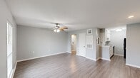 Located at 3400 Garden Oaks Dr in New Orleans, LA, this single-floor unit offers a well-organized layout comprising a living room, hallway, bathroom, and closet, with a kitchen visible from the main living area. The living room is the centerpiece of the space, finished with smooth light gray laminate flooring and neutral gray walls that lend a clean, contemporary feel. A ceiling fan with an integrated light fixture is centrally positioned, providing both airflow and ambient lighting. Natural light enters through a window fitted with horizontal blinds, brightening the room. The kitchen is accessible from the main living area and features white cabinetry and black appliances, including a refrigerator and range, offering a functional cooking space. A hallway connects the living area to the bathroom and closet, maintaining an efficient flow throughout. The bathroom includes a full soaking tub and shower combination, surrounded by large-format beige ceramic wall tiles, complemented by chrome fixtures and a standard toilet. A dedicated closet provides practical storage. White baseboards and trim throughout the unit add a crisp finishing touch, tying together the cohesive, neutral aesthetic of this straightforward and functional New Orleans residence.