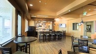 This single-floor bar and lounge space exudes a warm, casual ambiance, enhanced by rich earth-toned walls in a deep terracotta hue that create an inviting atmosphere throughout. The centerpiece of the space is a well-appointed full-service bar, framed by a handsome wooden archway and backlit shelving displaying a curated selection of spirits and glassware. A row of dark wood bar stools lines the bar counter, offering ample seating for guests. Pendant lighting with dark metal fixtures hangs above, complementing the recessed ceiling lights that illuminate the space evenly. The flooring features warm hardwood-style planks that tie the room together cohesively. Beyond the bar, additional seating areas with dark wood tables and chairs provide versatile arrangements for guests. A flat-screen television is mounted on the wall, adding an entertainment element to the lounge. Natural light filters in through windows fitted with horizontal blinds, while a full-size refrigerator positioned near the bar ensures convenient beverage service. The overall layout balances functionality and comfort, making it well-suited for social gatherings and relaxed dining experiences.