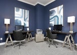 This single-story workspace is defined by its bold, deep navy walls that create a rich and immersive atmosphere conducive to focus and productivity. The room is finished with crisp white crown molding, providing an elegant contrast that frames the space with a refined, polished look. A flush-mount drum ceiling light with a fabric shade serves as the central overhead fixture, casting a warm, even glow throughout the room.

Two dedicated workstations are positioned along adjacent walls, each featuring sleek dark desks supported by trestle-style legs, paired with ergonomic black task chairs on casters. Each station is equipped with a desktop computer monitor, accommodating multiple users simultaneously. A stylish table lamp with a chrome base and white shade adds supplemental task lighting at one of the workstations.

The walls are adorned with a pair of large-scale abstract artworks in coordinating tones of navy, white, and gold, reinforcing the cohesive color palette. Neutral-toned carpet underfoot adds a layer of comfort and acoustic softness to the space. A cube-style side table in a textured finish sits centrally, offering additional surface space. The overall ambiance is sleek, modern, and purposefully designed.