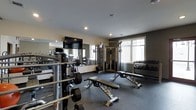 This fitness center is designed to support a comprehensive workout experience within a single, thoughtfully organized floor. The space is outfitted with a robust selection of strength training equipment, including a multi-function cable machine, a full dumbbell rack with a wide range of weights, and multiple adjustable workout benches suited for a variety of exercises. Free weights and barbells are readily accessible, catering to both beginner and advanced training needs. A large wall mirror spans one side of the room, enhancing the sense of space while allowing users to monitor their form during exercise. A wall-mounted flat-screen television is positioned prominently above the mirror, providing entertainment during workouts. The flooring features dark, durable fitness matting appropriate for heavy equipment and high-traffic use. Recessed lighting in the smooth ceiling keeps the space well-lit, complemented by natural light streaming in through multiple windows along the exterior wall. The neutral color palette of light and charcoal gray walls contributes to a clean, focused atmosphere. Overall, the fitness center strikes a balance between functionality and comfort, making it a well-rounded facility for strength and conditioning training.