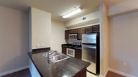 This single-floor residential space is highlighted by a well-appointed kitchen that serves as its centerpiece. The kitchen is designed for both functionality and style, featuring rich dark granite countertops that contrast elegantly with the dark espresso-toned cabinetry. A suite of stainless steel appliances — including a refrigerator, electric range, built-in microwave, and dishwasher — provides a modern, cohesive aesthetic. The double-basin stainless steel sink is thoughtfully positioned on a peninsula-style counter, offering additional workspace and a natural dividing point within the kitchen layout. Light tile flooring runs throughout, lending a clean, neutral foundation that complements the darker finishes above. The kitchen is illuminated by overhead fluorescent lighting, ensuring a bright and practical workspace. The walls are finished in a warm neutral tone, creating a welcoming ambiance throughout. The galley-style layout maximizes the use of available space, with cabinetry lining both sides to provide ample storage. Overall, the space presents a tidy, modern interior that balances practical design with contemporary residential finishes.