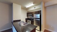 This single-floor residential space is highlighted by a well-appointed kitchen that serves as its centerpiece. The kitchen is designed for both functionality and style, featuring rich dark granite countertops that contrast elegantly with the dark espresso-toned cabinetry. A suite of stainless steel appliances — including a refrigerator, electric range, built-in microwave, and dishwasher — provides a modern, cohesive aesthetic. The double-basin stainless steel sink is thoughtfully positioned on a peninsula-style counter, offering additional workspace and a natural dividing point within the kitchen layout. Light tile flooring runs throughout, lending a clean, neutral foundation that complements the darker finishes above. The kitchen is illuminated by overhead fluorescent lighting, ensuring a bright and practical workspace. The walls are finished in a warm neutral tone, creating a welcoming ambiance throughout. The galley-style layout maximizes the use of available space, with cabinetry lining both sides to provide ample storage. Overall, the space presents a tidy, modern interior that balances practical design with contemporary residential finishes.