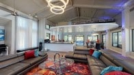 Located at 8100 US 98 in Pensacola, Florida, this single-story clubhouse is a well-appointed community gathering space that masterfully blends modern design with a welcoming, energetic atmosphere. The main living area serves as the heart of the facility, showcasing soaring vaulted ceilings with exposed structural elements and a dramatic triple-ring pendant light fixture. Rich hardwood-style flooring flows throughout, while a vibrant, large-format area rug anchors a spacious seating arrangement of leather sectional sofas accented with bold, colorful throw pillows.

A central kitchenette island with dark countertops and white cabinetry provides a functional refreshment hub, complete with beverage stations and ample counter space. A dedicated business center nook offers computer workstations and a wall-mounted display screen, framed by sheer floor-to-ceiling curtains that filter generous natural light.

Tall French doors open to a screened outdoor lounge that overlooks a tranquil waterfront setting surrounded by lush greenery. This covered patio features comfortable sectional seating, colorful area rugs, ceiling fans, and dining areas, creating an ideal space for relaxation or social gatherings. An additional open event room with soaring ceilings, ambient purple accent lighting, and expansive glass doors connects seamlessly to the outdoor pool area, completing this versatile and visually striking facility.