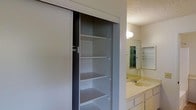 This single-floor space includes a bathroom that combines practicality with efficient use of space. A standout feature is the built-in linen closet fitted with sliding doors and multiple shelves, offering generous storage for towels, toiletries, and essentials. The vanity area features a light-toned countertop with an integrated sink, white cabinetry below, and a large mirror above, which helps reflect light and enhance the sense of space. A medicine cabinet with interior shelving is also visible, providing additional organized storage. The walls have a smooth finish, while the ceiling features a textured popcorn surface typical of the construction era. Wall-mounted lighting above the mirror provides focused illumination for the vanity area. The overall design is straightforward and utilitarian, prioritizing storage and functionality. Neutral tones throughout — including beige countertops, white cabinetry, and gray-toned closet interiors — contribute to a clean, understated aesthetic. The layout makes efficient use of the available space, with storage solutions integrated directly into the bathroom's design.