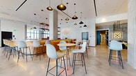 Located at 2126 Arapahoe St in Denver, Colorado, this single-floor amenity space showcases a thoughtfully designed interior that blends industrial and modern aesthetics. Polished concrete floors extend throughout, anchoring an open, flowing layout that transitions seamlessly between distinct functional zones.

At the heart of the space sits a curved café-style bar clad in warm wood tile, backlit for ambiance and lined with sleek white counter-height stools. Behind it, a bold red open-shelf display unit adds a striking contrast to the surrounding neutral palette. Clusters of round bistro tables paired with contemporary shell-style bar chairs offer casual seating throughout the dining area, while a curated collection of pendant lights—ranging from sculptural black dome fixtures to exposed-bulb geometric arrangements—creates visual interest overhead.

The lounge areas feature plush seating in muted greens, blues, and mustard tones arranged around low wood coffee tables atop natural woven rugs. Floor-to-ceiling glass curtain walls flood these areas with abundant natural light and frame panoramic views of the Denver skyline and an adjacent outdoor terrace. A boldly painted concrete accent wall with teal and red geometric artwork adds a distinctive artistic focal point to one lounge section, reinforcing the space's modern, urban character.