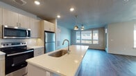 This contemporary single-floor residence offers a thoughtfully designed open-concept layout that seamlessly connects the kitchen, dining, and living areas. The kitchen is a standout feature, showcasing light wood cabinetry extending to the ceiling for generous storage, white quartz countertops, and a full suite of stainless steel appliances including a refrigerator, electric range, and over-the-range microwave. A central island with an undermount sink and pendant lighting above serves as both a functional workspace and a natural gathering point.

Rich hardwood-style flooring flows throughout the main living areas, adding warmth and continuity to the space. The open living area is bright and airy, benefiting from multiple windows that invite natural light and offer outdoor views. Recessed lighting and additional pendant fixtures provide layered illumination throughout.

The bathroom is efficiently designed, featuring a walk-in shower with a sliding glass door, a vanity with storage, and hardwood-style flooring consistent with the rest of the unit. A stacked washer and dryer unit is also conveniently located within the space. The overall design palette—neutral walls, light cabinetry, and warm flooring tones—creates a clean, modern ambiance that is both functional and welcoming.