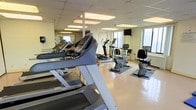 This fitness center in Silver Spring, Maryland, offers a functional and well-maintained exercise environment designed to support a range of cardio-focused workouts. The space is outfitted with a selection of professional-grade Precor cardio equipment, including treadmills, elliptical trainers, and stationary bikes, arranged to allow comfortable movement throughout the room. Rubber floor mats are positioned beneath the equipment, providing stability and floor protection. A wall of mirrors along one side of the room visually expands the space and allows users to monitor their form during workouts. Large windows on the opposite wall admit natural light, contributing to an open and airy atmosphere while offering views of the surrounding area. A wall-mounted television is also present, providing entertainment options during exercise sessions. The dropped tile ceiling features recessed panel lighting that ensures the space remains well-lit throughout the day. The light-toned flooring and neutral walls create a clean, practical environment. Overall, the fitness center presents a straightforward and well-organized layout suited for daily cardio training.