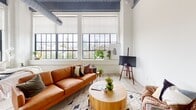 Located at 2501 Oakford Street in Philadelphia's Point Breeze neighborhood, this single-floor unit showcases an industrial-loft aesthetic that blends raw architectural elements with contemporary design. The space is defined by its dramatic ceiling height, where exposed ductwork, steel beams, and a striking navy-painted wood ceiling create a bold overhead statement. Light hardwood flooring runs throughout, complementing the crisp white walls and lending warmth to the industrial framework.

The open living area is anchored by oversized factory-style windows with black steel frames, flooding the space with abundant natural light and offering expansive views of the Philadelphia skyline. The kitchen is sleek and functional, outfitted with white cabinetry, subway tile backsplash, stainless steel appliances — including a full-size refrigerator, range, dishwasher, and microwave — and light countertops. A large steel-framed window nearby brings in additional natural light and treetop views.

The bathroom is finished with clean, modern details, including a frameless glass-enclosed shower with white tile surround, a floating vanity, and neutral tile flooring. Throughout the unit, the interplay of industrial structure and refined finishes creates a cohesive, urban ambiance that is both functional and visually compelling.