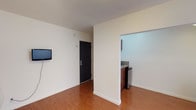 This single-floor unit offers a practical and efficiently designed layout with warm hardwood-style flooring running throughout the main living areas. The open living space features neutral white walls, a wall-mounted flat-screen TV, and a dark entry door, creating a clean and functional environment. A dedicated kitchenette is accessible via an open archway and includes rich dark wood cabinetry, granite-look countertops, a stainless steel single-basin sink, a two-burner gas cooktop, a mounted microwave, and a compact refrigerator — providing essential cooking and storage functionality in a well-organized footprint. A separate bedroom area benefits from natural light streaming through a large window fitted with vertical blinds, complemented by a flush-mount ceiling light fixture. The bathroom features a white pedestal-style sink, a mirrored medicine cabinet for storage, and tile flooring. Throughout the unit, crisp white walls, consistent flooring materials, and practical fixtures contribute to a tidy, no-frills ambiance suitable for everyday living.