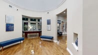 This thoughtfully designed single-floor space offers a generous layout across its various functional areas. The architectural centerpiece is a dramatic rotunda-style entrance foyer, distinguished by a sweeping curved ceiling with detailed crown molding and polished hardwood floors that reflect natural light. Symmetrical wall niches flank a set of stately wood-framed French doors, creating a refined first impression.

The primary living area showcases a domed, curved ceiling and flows openly into a game room featuring a billiard table and expansive windows that draw in abundant natural light. A secondary living area centers around a striking fireplace with a white decorative mantel set against a bold navy accent wall, complemented by a wall-mounted television and large windows.

The hallway connects the various spaces seamlessly, passing an office area with a frosted-glass branded panel and dedicated workspace. The kitchen is well-equipped with rich dark wood cabinetry, granite countertops, a herringbone tile backsplash, and tile flooring. An additional room provides a quiet, intimate seating area with natural light. Throughout, warm hardwood floors, neutral tones, crown molding, and arched doorways contribute to a cohesive and welcoming atmosphere.