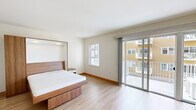 This thoughtfully designed single-floor unit offers a clean, contemporary aesthetic throughout, characterized by light hardwood flooring, crisp white walls, and an abundance of natural light. The open-concept layout connects the living and kitchen areas seamlessly, creating an airy and functional flow.

The kitchen is a standout feature, outfitted with sleek white cabinetry extending to the ceiling, dark granite countertops, and a full suite of stainless steel appliances including a refrigerator, dishwasher, oven, and built-in microwave. Recessed lighting illuminates the space with precision, complementing the modern, minimalist design.

The bedroom is generously proportioned, offering a warm, restful atmosphere with light wood flooring and a large sliding glass door that opens directly onto a private balcony, allowing for seamless indoor-outdoor living and views of the surrounding building community.

The living area benefits from its own sliding glass door to a second outdoor balcony and is equipped with a wall-mounted mini-split HVAC unit, ensuring year-round comfort. The bathroom features a dark granite vanity countertop, a wide mirror, and warm wood-trimmed doorframes that echo the natural tones found throughout the unit. Overall, the space exudes a fresh, modern character with functional, high-quality finishes.