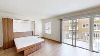 This thoughtfully designed single-floor unit offers a clean, contemporary aesthetic throughout, characterized by light hardwood flooring, crisp white walls, and an abundance of natural light. The open-concept layout connects the living and kitchen areas seamlessly, creating an airy and functional flow.

The kitchen is a standout feature, outfitted with sleek white cabinetry extending to the ceiling, dark granite countertops, and a full suite of stainless steel appliances including a refrigerator, dishwasher, oven, and built-in microwave. Recessed lighting illuminates the space with precision, complementing the modern, minimalist design.

The bedroom is generously proportioned, offering a warm, restful atmosphere with light wood flooring and a large sliding glass door that opens directly onto a private balcony, allowing for seamless indoor-outdoor living and views of the surrounding building community.

The living area benefits from its own sliding glass door to a second outdoor balcony and is equipped with a wall-mounted mini-split HVAC unit, ensuring year-round comfort. The bathroom features a dark granite vanity countertop, a wide mirror, and warm wood-trimmed doorframes that echo the natural tones found throughout the unit. Overall, the space exudes a fresh, modern character with functional, high-quality finishes.