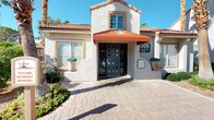 This apartment community showcases a distinctive Mediterranean-inspired architectural style, characterized by terracotta tile rooflines, stucco exteriors, and warm earth-tone finishes throughout. The welcoming entrance features a paved courtyard approach, decorative awning, and manicured landscaping that sets the tone for the community's polished aesthetic.

The outdoor spaces are a standout feature of this property. A generously sized patio area incorporates multiple social zones, beginning with a striking stacked-stone fireplace flanked by cushioned lounge seating — ideal for relaxed gatherings. Nearby, a substantial outdoor kitchen station provides stainless steel appliances, a built-in grill, refrigerator, and storage, accompanied by a large bar-height counter with multiple bar stools and striped shade umbrellas overhead.

A beautifully crafted wood-beamed gazebo serves as a sheltered lounge area, furnished with patterned sofas and a coffee table atop a decorative area rug, overlooking a serene courtyard fountain. Mature palm trees and lush greenery frame the outdoor spaces throughout, providing shade and a resort-like ambiance beneath bright, sunny skies.
