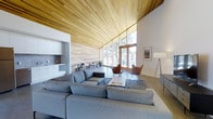This single-floor space is defined by its exceptional architectural character and thoughtful design. The most striking feature is the sweeping vaulted ceiling clad in light natural wood planks, which rises dramatically and draws the eye toward an expansive wall of floor-to-ceiling windows. These large glazed panels flood the interior with abundant natural light and offer open views of the surrounding trees and neighboring structures, creating a seamless connection between indoors and outdoors.

The open-plan layout accommodates several distinct functional zones. The main lounge area is anchored by a generous sectional sofa in soft grey, paired with a minimalist coffee table and mid-century-inspired leather butterfly chairs, lending the space a modern yet comfortable character. Adjacent to the lounge, a dining area features a high table and chairs positioned near the windows, making it an inviting spot to gather.

A compact kitchenette is integrated along one wall, outfitted with white cabinetry, subway tile backsplash, and a countertop workspace, providing practical amenities without disrupting the open flow of the space. A natural wood accent wall adds warmth and texture, complementing the ceiling's palette.

Overall, the space strikes a balance between contemporary minimalism and natural warmth, making it a versatile and visually engaging environment.