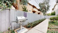 Located at 10740 Blix St in the North Hollywood neighborhood of Los Angeles, this outdoor common area offers residents a well-appointed shared space designed for relaxation and social gatherings. The grounds are anchored by a stainless steel built-in barbecue grill station, complete with posted community barbecue rules, making it a practical and inviting spot for outdoor cooking and entertaining. Adjacent to the grill, a spacious concrete patio accommodates a wooden dining table and cushioned chairs, providing a comfortable setting for al fresco dining. The courtyard is enclosed by a gray wood privacy fence that defines the space and creates a sense of seclusion. Lush ground cover plantings and mature trees, including a prominent pine, line the perimeter walkways and landscaped beds, adding greenery and natural character to the environment. Wide concrete pathways connect the various areas of the grounds, offering easy circulation throughout. The multi-story stucco residential building frames the space, with balconies visible above and covered parking accessed via a gated area at ground level. The overall ambiance is functional and community-oriented, with thoughtful landscaping and amenities that encourage outdoor living in the mild Southern California climate.