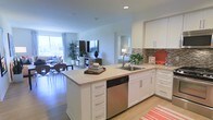 This single-floor residence showcases a thoughtfully designed open-concept layout that integrates the kitchen, dining, and living areas into one cohesive, light-filled space. The kitchen is a central highlight, featuring crisp white cabinetry, light stone countertops, and a peninsula island with built-in drawers that provides additional workspace and casual seating potential. A stainless steel dishwasher and undermount sink add a polished, functional touch, while a tile backsplash in earthy tones introduces a subtle textural contrast. The kitchen transitions naturally into the dining area, where a large window allows generous natural light to fill the room, creating an airy, welcoming ambiance. The living area extends further, offering a comfortable entertainment setup and views to the outdoors through expansive windows. Light hardwood flooring runs throughout, unifying the open space while contributing to its clean, contemporary aesthetic. Glimpses of an adjoining room suggest a well-connected floor plan. The overall atmosphere is modern, bright, and functional, well-suited for everyday living and entertaining.