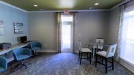 This single-floor space presents a functional and well-appointed interior designed for casual gatherings or meetings. The room features neutral gray walls accented by clean white crown molding and baseboards, creating a polished, cohesive aesthetic throughout. Recessed lighting in the ceiling provides consistent overhead illumination, complementing the natural light that filters through a glazed exit door framed by floor-length patterned drapes. The door's glass panels allow visibility to the outdoors while maintaining a sense of enclosure. The flooring consists of a textured, multi-toned patterned carpet that adds visual interest and a degree of warmth underfoot. A round glass-top dining table with high-backed upholstered chairs occupies one side of the room, offering a practical surface for work or dining. On the opposite side, a cushioned blue accent chair with a swivel base provides a relaxed seating option. The overall ambiance is professional yet approachable, blending functional furnishings with thoughtful design details that make the space suitable for both social and work-oriented activities.