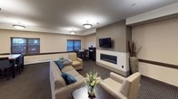 This single-floor community space is thoughtfully designed to serve as a comfortable and functional gathering area. The layout is divided into distinct zones that flow seamlessly into one another, creating an inviting and versatile environment.

The lounge area serves as the centerpiece of the space, anchored by a sleek, wall-mounted linear fireplace set against a large tile surround. A flat-screen television is mounted above, making it ideal for relaxed viewing. Comfortable seating, including a plush sofa accented with blue throw pillows and cream-toned armchairs, is arranged to encourage conversation and community gathering. Warm pendant lighting and flush-mount ceiling fixtures illuminate the space throughout.

The fully equipped kitchen features rich dark-wood cabinetry with granite countertops, stainless steel appliances including a full-size refrigerator, a gas range with oven, and a microwave. A large island with bar seating provides a casual space for dining or socializing. Tile flooring in the kitchen transitions to patterned carpet in the lounge and dining areas.

Multiple dining tables with dark chairs are positioned near windows dressed with wood-toned blinds, allowing natural light to filter in. Neutral beige walls accented with dark wood trim tie the entire space together, lending it a warm, sophisticated ambiance suited for shared community use.