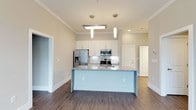 This single-floor space showcases a thoughtfully designed open-concept layout centered around a modern kitchen. The kitchen serves as the focal point, featuring a large central island topped with speckled granite countertops that provide ample workspace and a natural gathering spot. The island's clean white base complements the matching white shaker-style cabinetry that lines the walls, offering generous storage throughout.

Stainless steel appliances, including a French door refrigerator with ice and water dispenser, a built-in microwave, and a freestanding range, add a polished, contemporary feel. The kitchen is illuminated by a combination of recessed lighting, a flush-mount ceiling fixture, and elegant cylindrical pendant lights that hang gracefully above the island.

Crown molding runs along the ceiling perimeter, adding a refined architectural detail to the space. The warm-toned hardwood-style flooring extends throughout, providing a cohesive and inviting aesthetic. Neutral beige walls keep the space bright and versatile. A doorway visible in the background suggests seamless flow into additional rooms, reinforcing the open and connected nature of the overall floor plan.