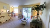 This 2-floor residence offers a welcoming and airy atmosphere, highlighted by warm yellow walls that lend a sunny, cheerful character throughout the living spaces. The main living area is generously proportioned, featuring soft carpeted flooring and a ceiling fan that contributes to a comfortable environment. A prominent sliding glass door floods the room with natural light and opens onto a private balcony, where greenery and garden views create a pleasant indoor-outdoor connection framed by white curtains with coral accents.

The living area is thoughtfully laid out to accommodate both relaxed seating and casual entertaining, with space for a wicker-style coffee table and coordinating furniture. Adjacent to the living room, a dedicated dining area is visible, featuring a dining table and chairs positioned near a window with vertical blinds, allowing for additional natural light. Decorative touches, including tropical potted plants and framed wall art, enhance the warm, inviting ambiance. The overall design of the space balances functionality with a light, tropical-inspired aesthetic that feels both comfortable and livable across its two floors.